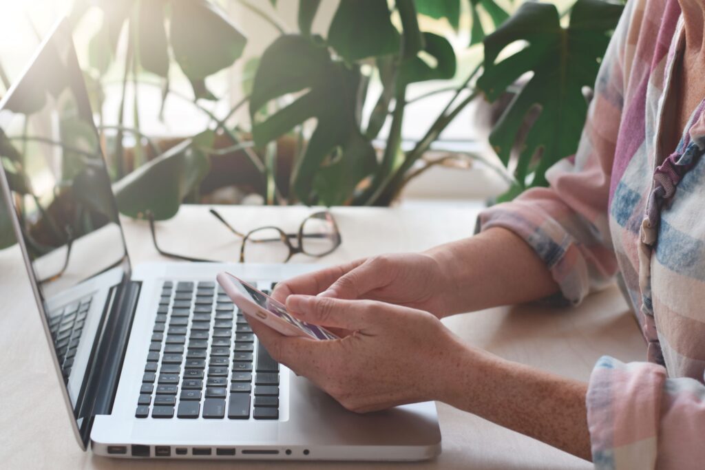 A woman uses her smartphone while sitting at a bright desk with an open laptop and a large monstera plant in the background.