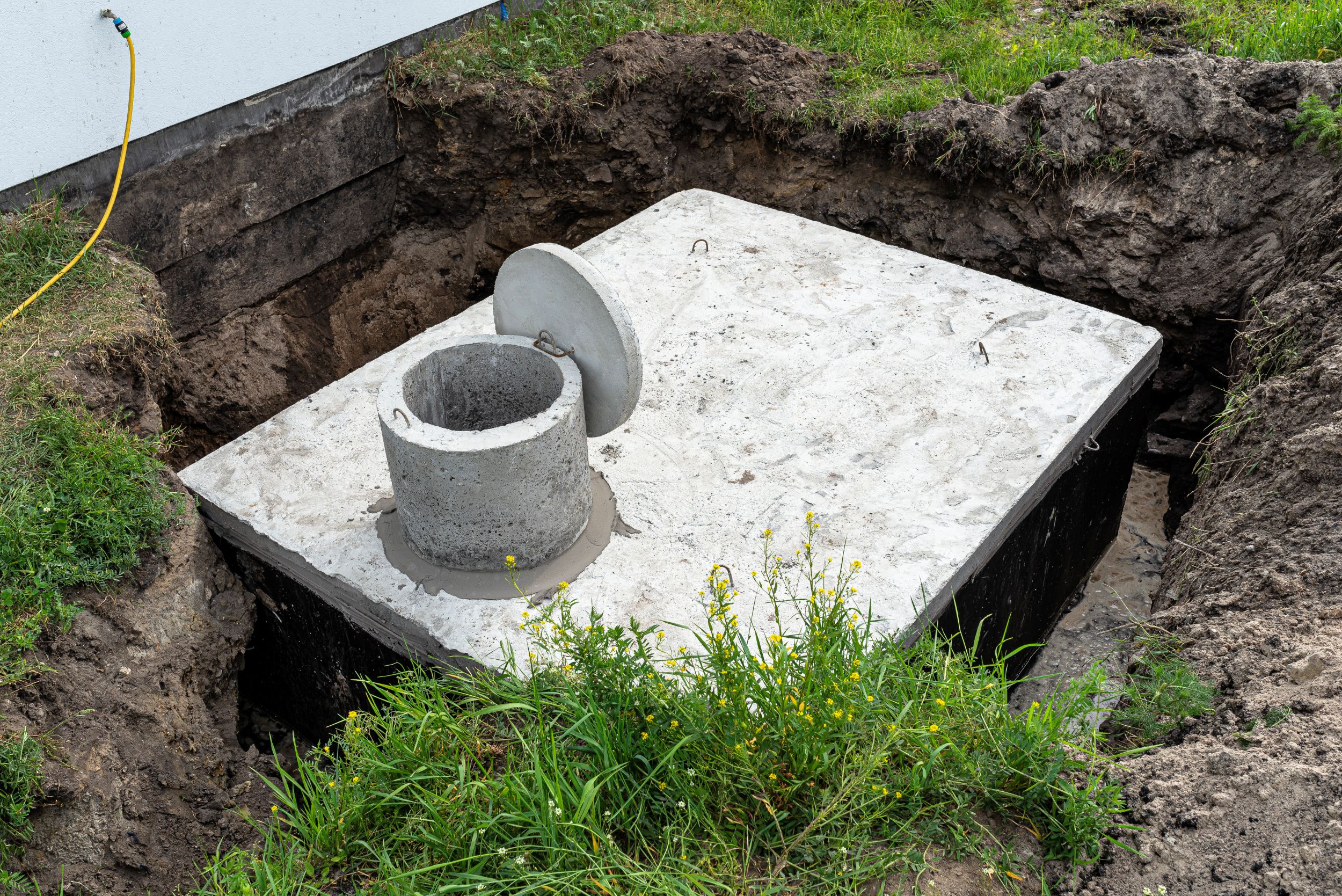 A rectangular concrete septic tank with a circular access riser and open lid sits in a dirt excavation pit next to a building foundation.