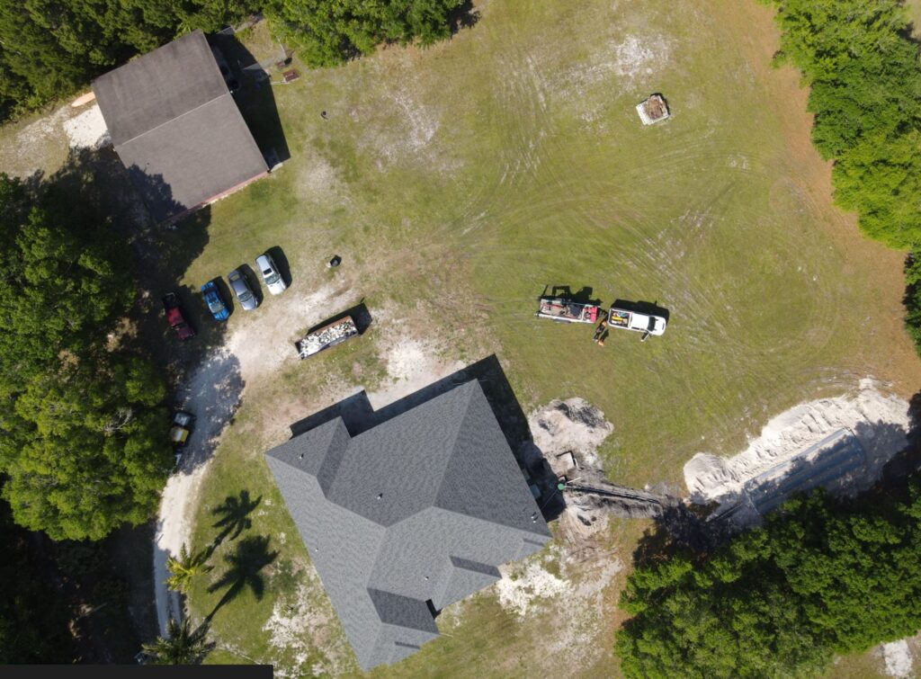An aerial drone view shows a service truck with a trailer parked on a lawn near a residential septic system installation, featuring a trench line running toward an excavated drain field.
