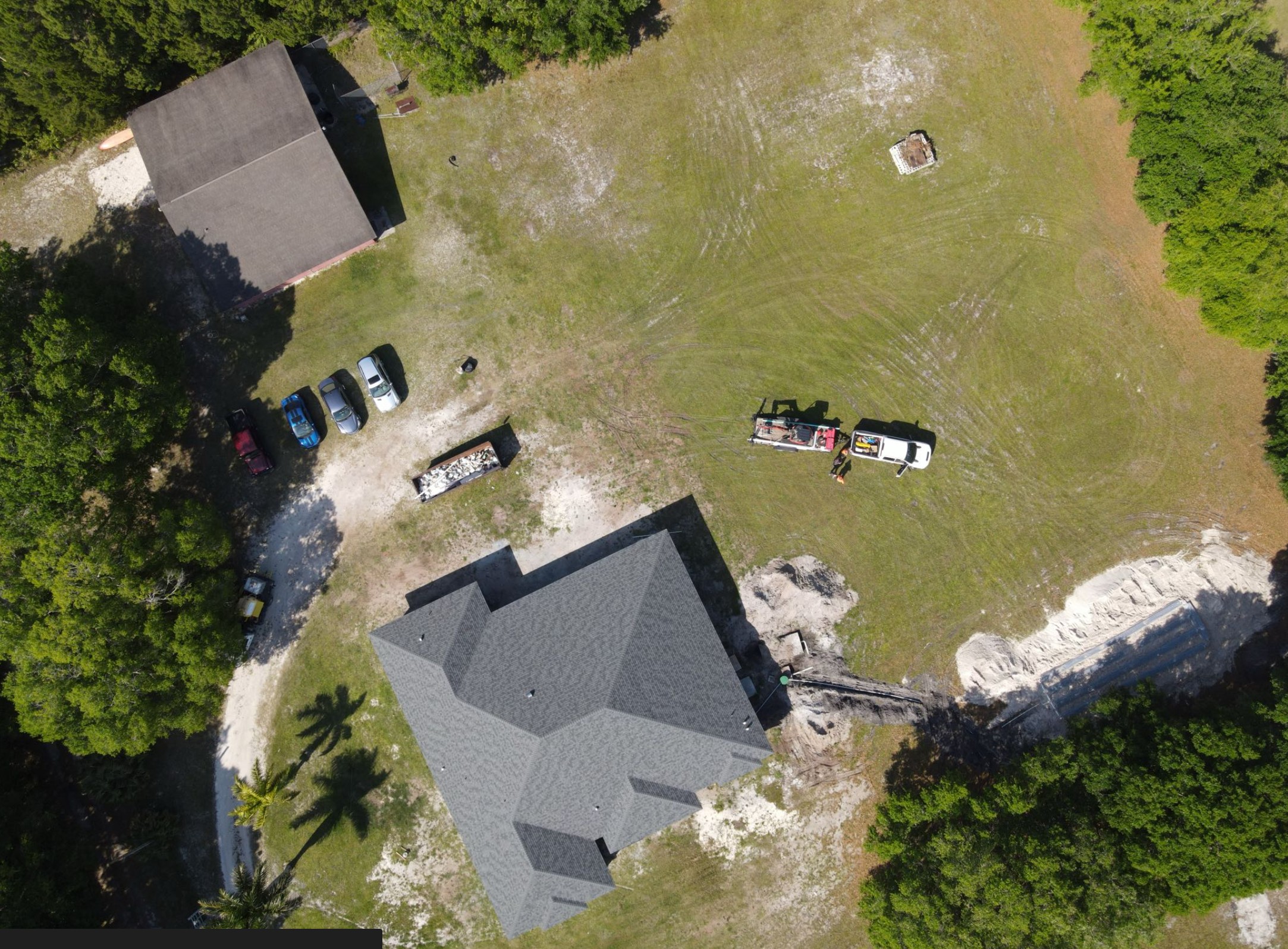 An aerial drone view shows a service truck with a trailer parked on a lawn near a residential septic system installation, featuring a trench line running toward an excavated drain field.