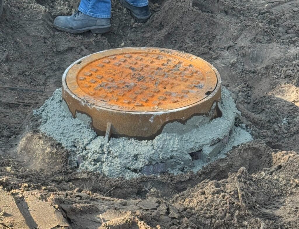 A round, rusted iron sanitary manhole cover is secured around its base with fresh concrete within an excavated dirt area.