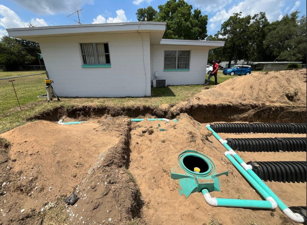 A residential septic system under construction features an open green tank riser, connected PVC piping, and parallel rows of black drain field chambers in an excavated backyard next to a white house.
