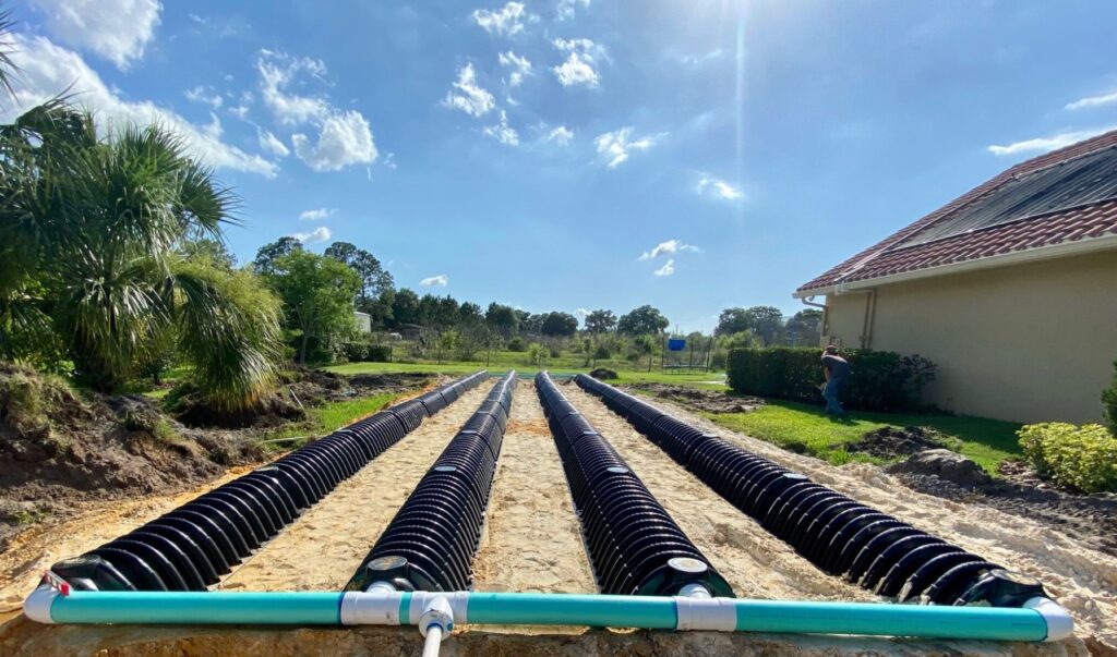 Four long, parallel rows of black corrugated septic drain field chambers are connected by a teal PVC manifold pipe in a residential backyard under construction.