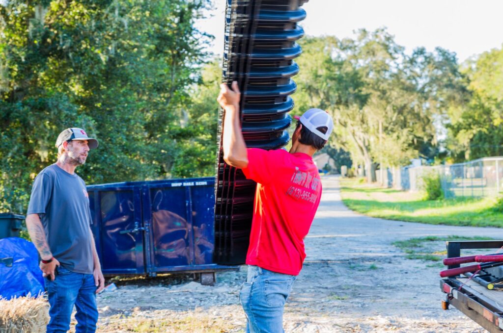 A Tampa Bay Septic technician in a red shirt lifts a stack of black corrugated drain field chambers while a coworker stands nearby at an outdoor job site.