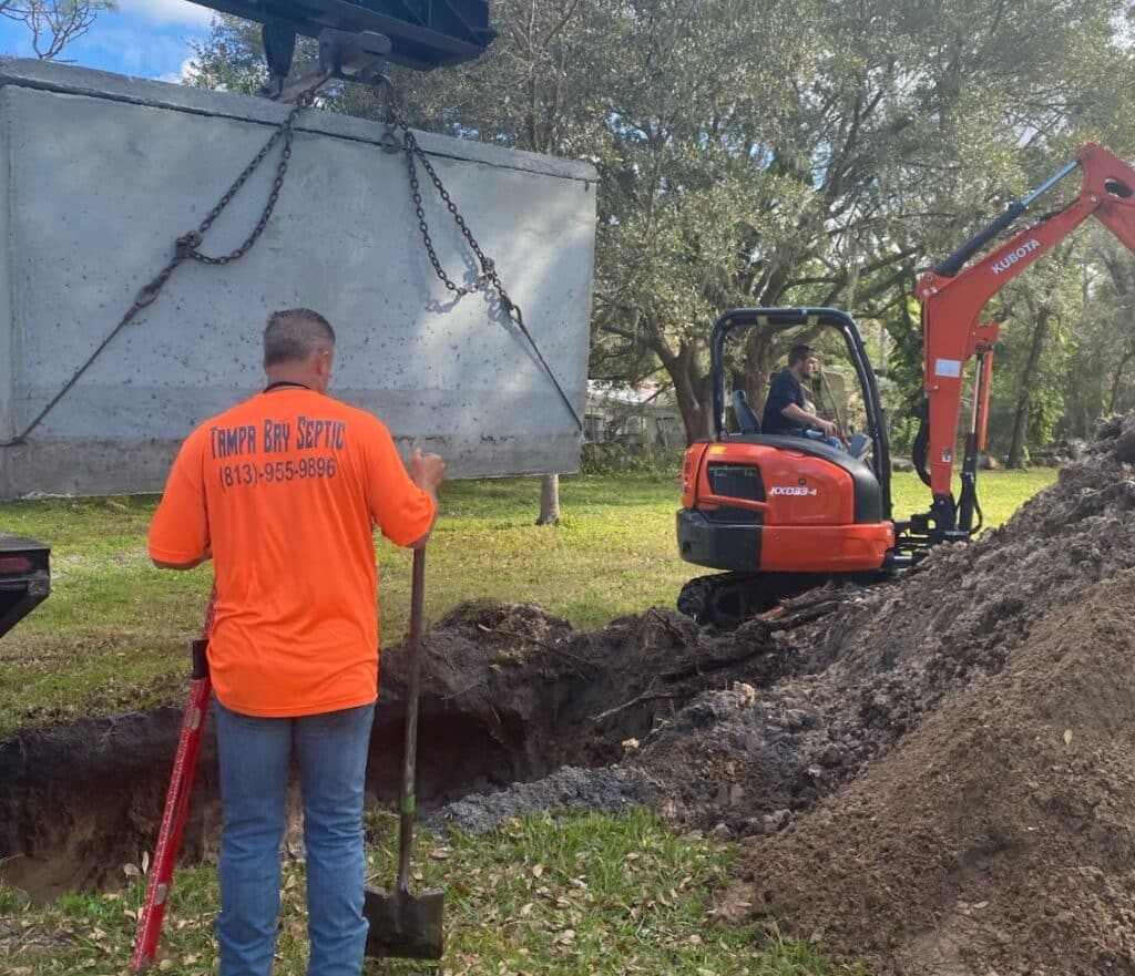A worker in a bright orange shirt watches a large precast concrete septic tank being hoisted by chains into an excavated pit during septic services in Orlando, FL, while an orange Kubota mini excavator sits nearby.