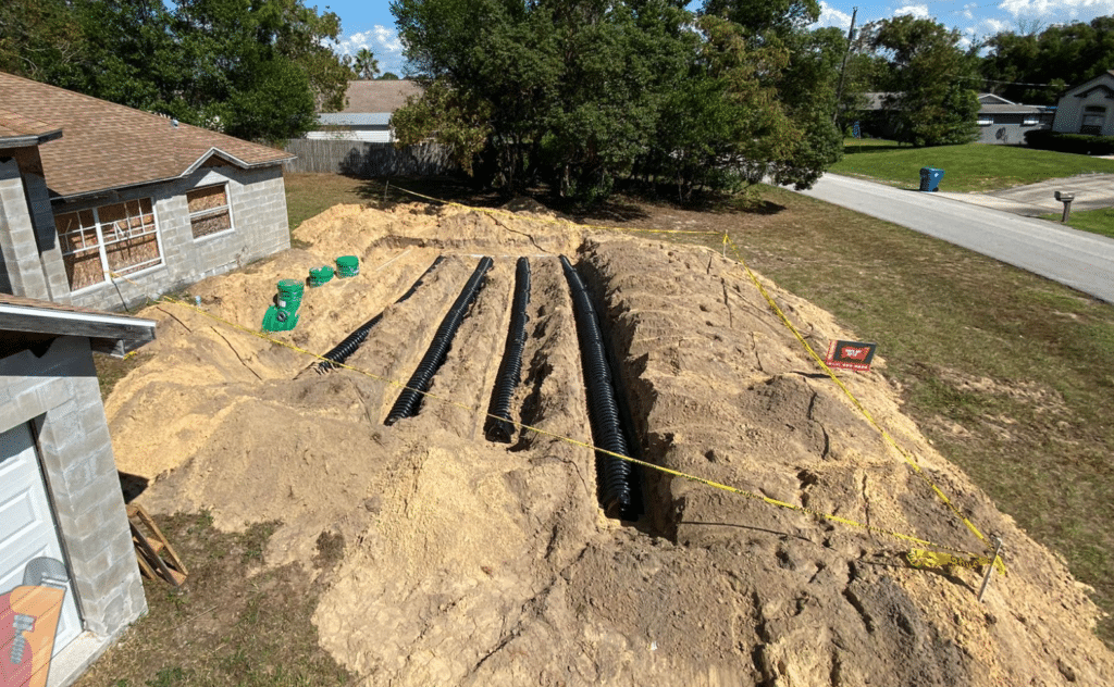 Four parallel rows of black corrugated plastic chambers are laid out in a large, elevated sand mound for a new septic system drainfield in a residential front yard, surrounded by yellow caution tape.