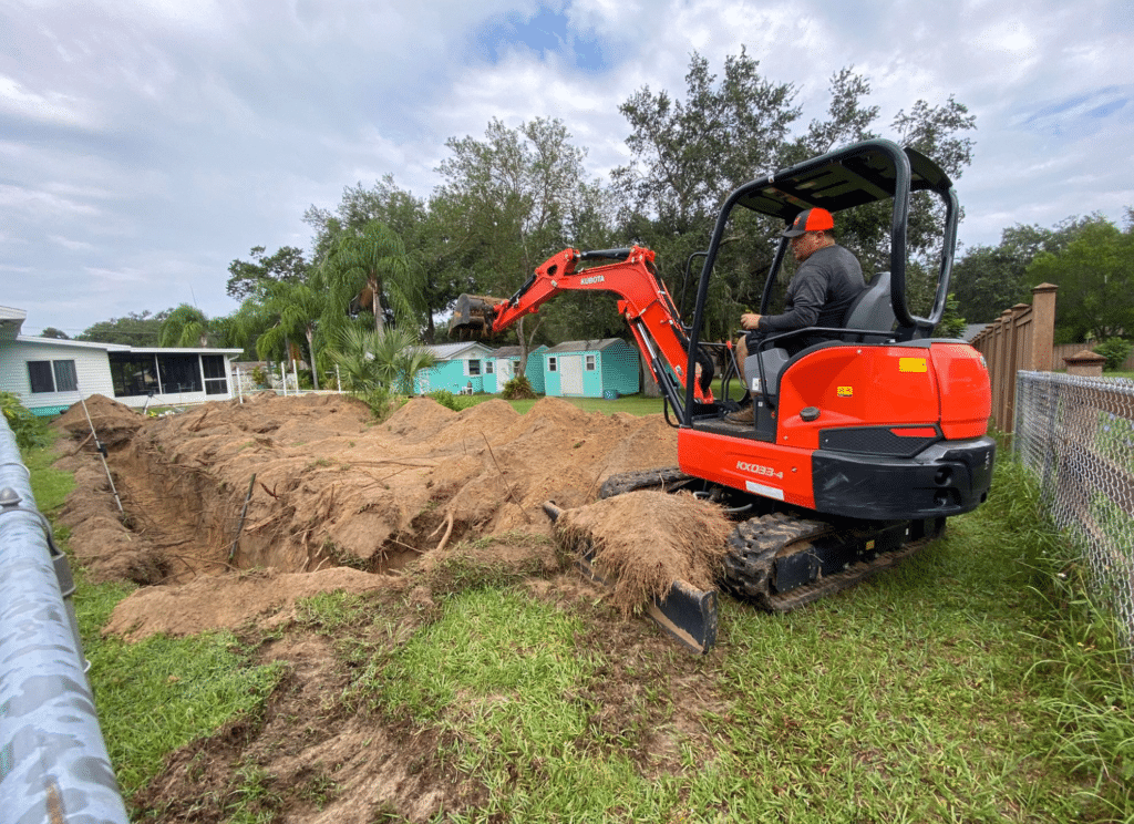 A man operates an orange Kubota mini excavator to dig a large trench in a residential backyard.