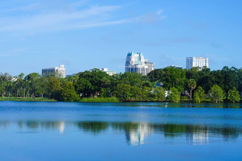 The downtown Orlando skyline rises behind a dense line of lush green trees, reflecting beautifully in the calm blue waters of a lake in the foreground.