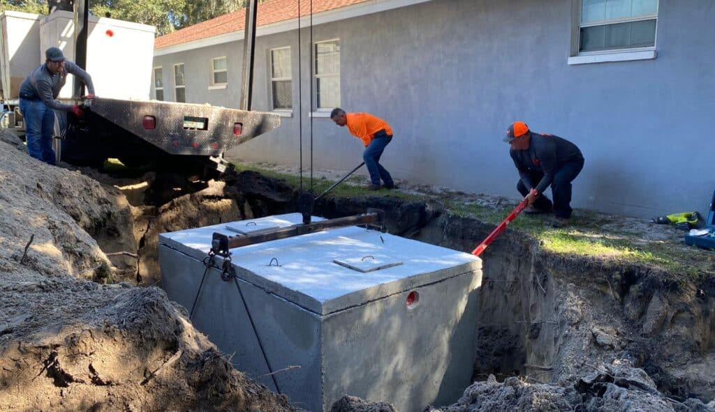 three construction workers guide a large precast concrete septic tank as it is lowered by cables into a deep excavated trench next to a house.
