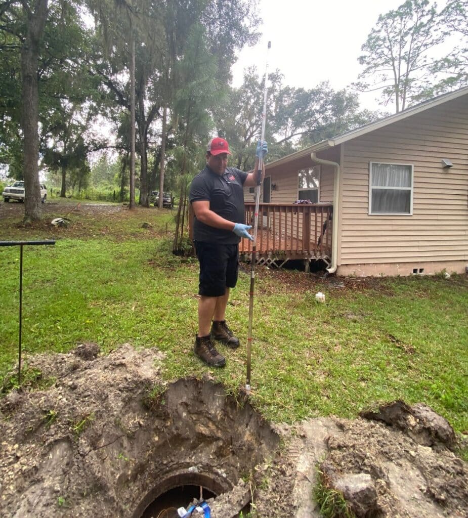 A service technician wearing a red cap and work boots using a long measuring rod to inspect a dug-up underground tank opening in a residential yard.