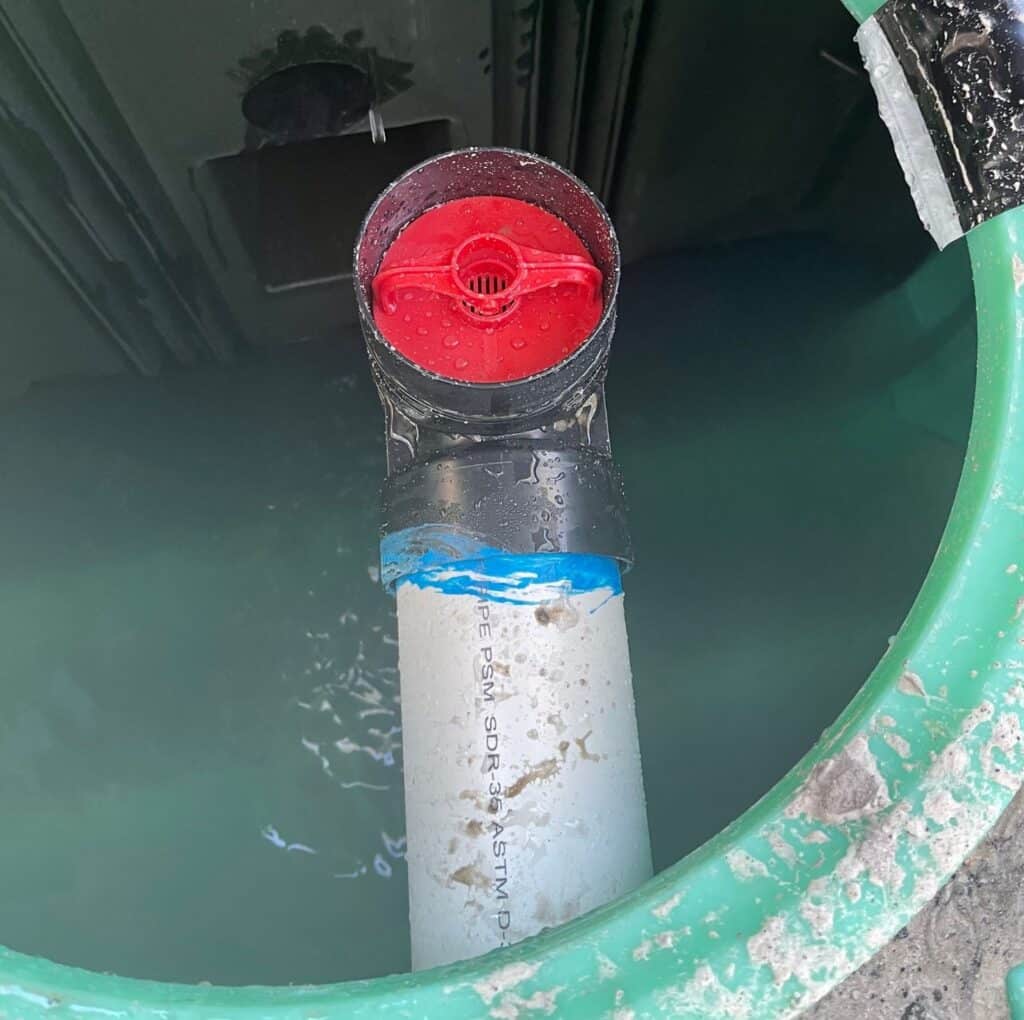 A top-down view inside a green septic tank riser filled with water shows a vertical white PVC pipe topped with a black fitting and a bright red effluent filter cap.