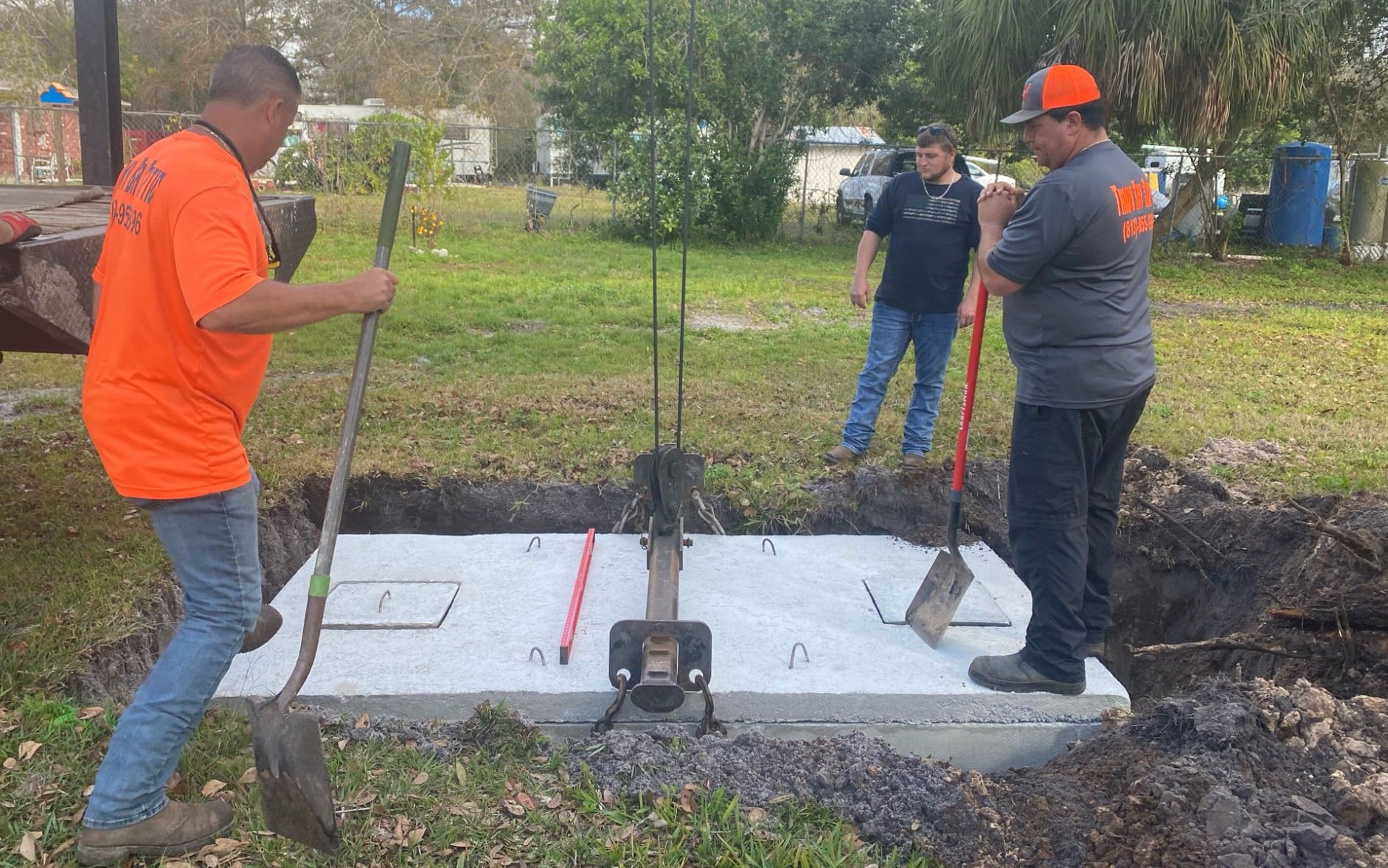 Three workers use shovels to install a large concrete septic tank lid into an excavated pit in a grassy yard.