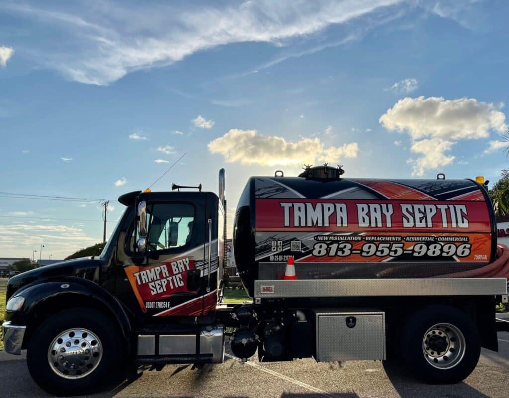 A black commercial septic truck with "Tampa Bay Septic" branding and a phone number on its large tank, parked outdoors under a bright, partly cloudy sky.