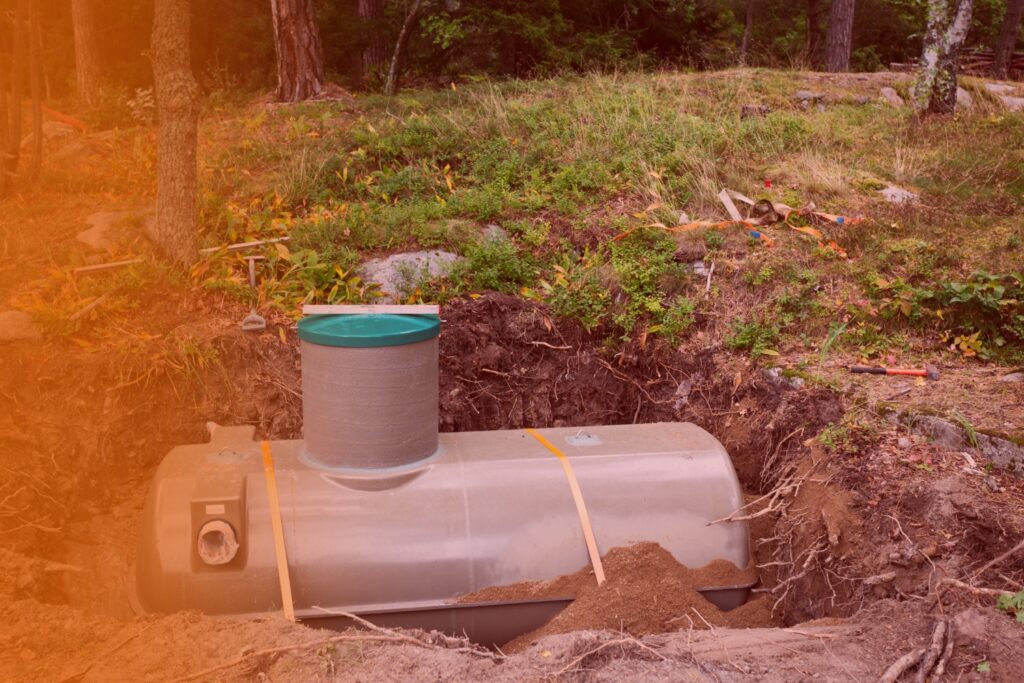 A large underground septic tank with a green riser pipe installed in an excavated dirt pit surrounded by trees and lawn.