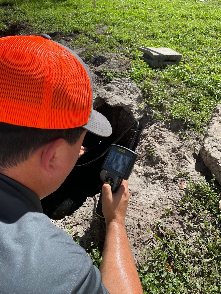 A worker wearing a bright orange cap uses a handheld digital inspection camera to peer into a dark opening in the ground.