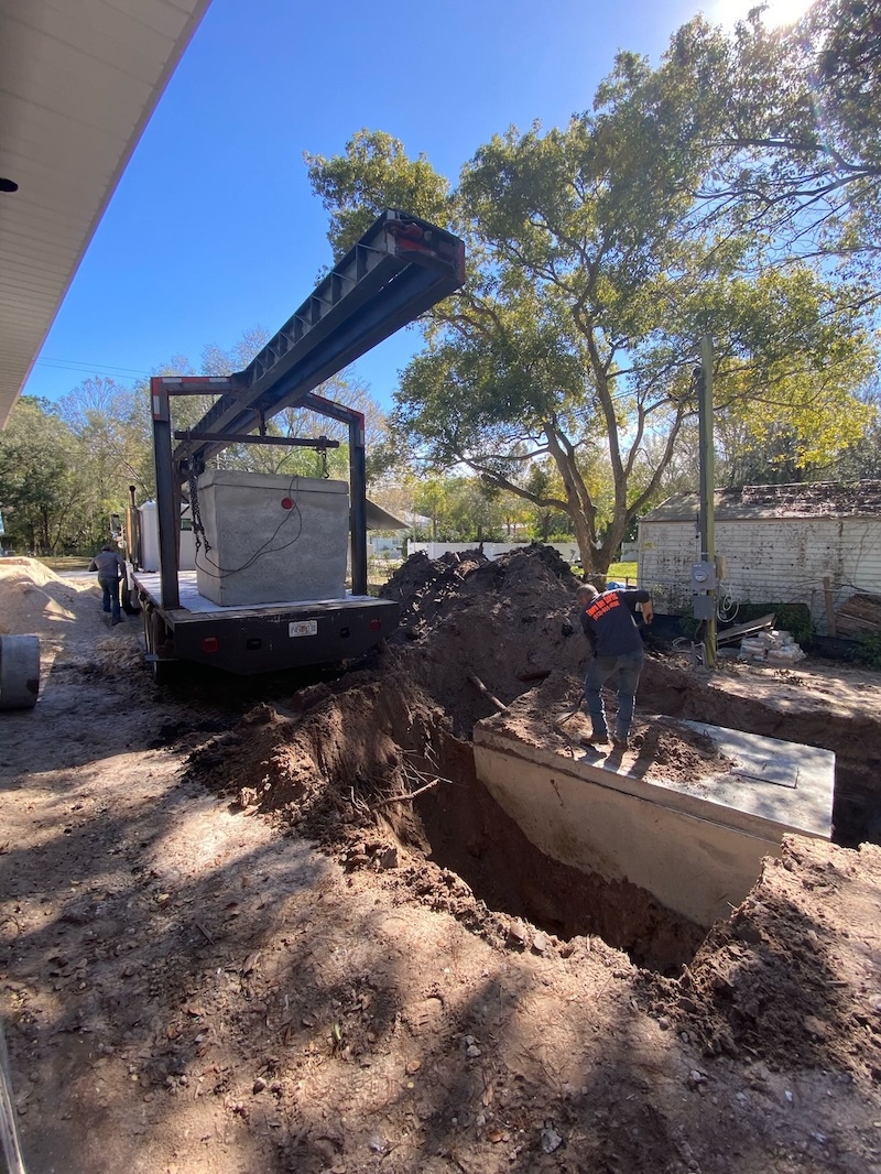 A worker stands on a concrete tank inside a deeply excavated trench while a boom truck positions another unit during a professional septic service fort myers fl.