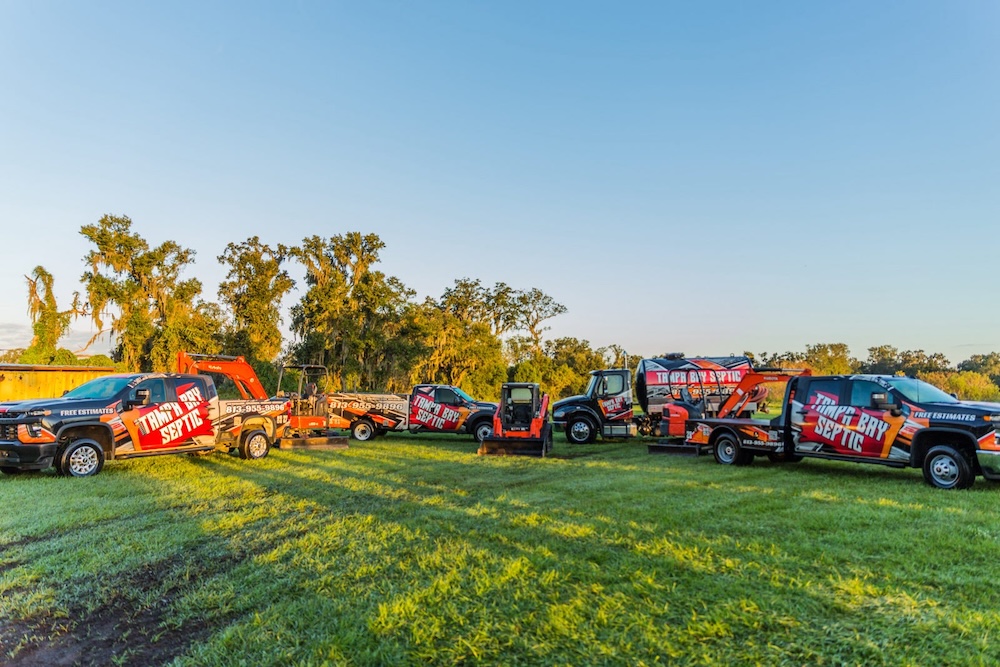 A fleet of "Tampa Bay Septic" commercial vehicles, including branded pickup trucks, a pumper truck, and excavation machinery, is parked in a row across a grassy field under a clear sky.