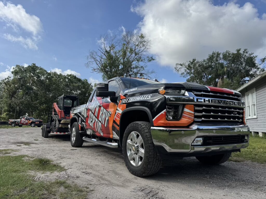A wrapped black, orange, and white Chevrolet pickup truck with "Tamp'N Bay Septic" branding is parked on a dirt path while towing a trailer carrying a red track loader.
