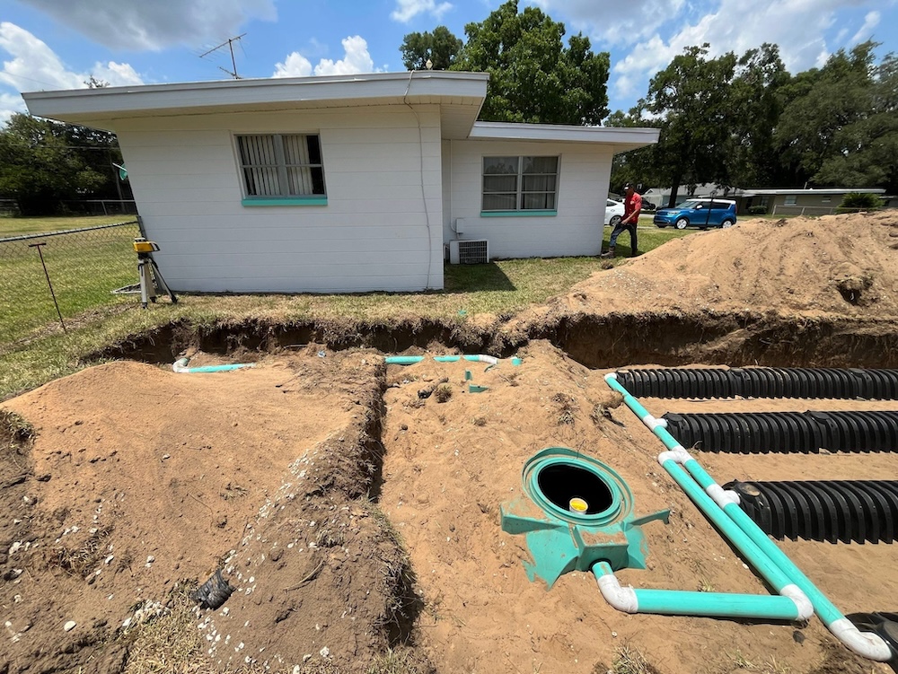 An excavated residential backyard features the installation of an underground septic system, displaying a green tank riser, PVC piping, and rows of black drainfield chambers behind a white, single-story house.