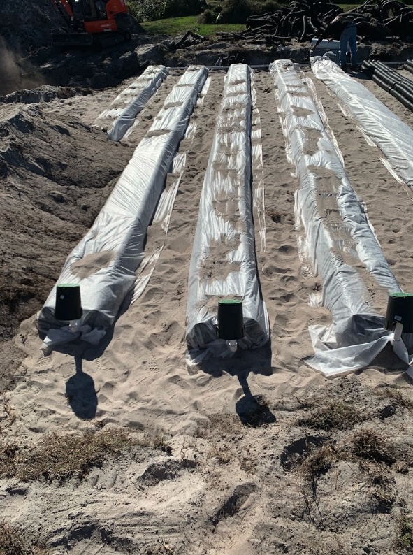 Parallel septic drain field pipes covered in protective fabric laid out in sand at a residential installation site.
