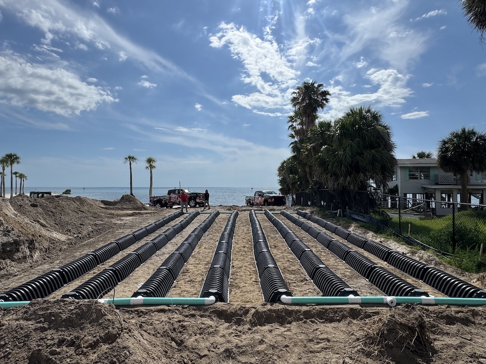 Parallel rows of black plastic septic drain field chambers installed in sandy ground on a waterfront property.