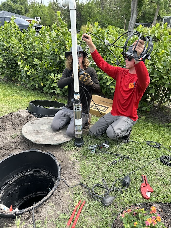 Two technicians kneel beside an open septic tank riser in a grassy lawn while wiring and preparing a new submersible pump for installation.