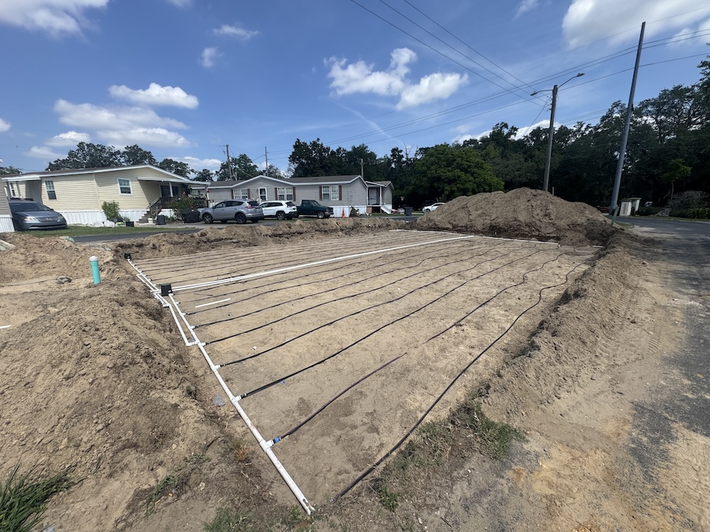 A low-pressure drip septic drain field featuring white PVC manifold pipes and black tubing laid out across a sandy installation bed in a residential neighborhood.