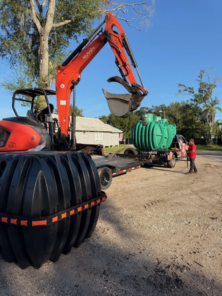 An orange Kubota excavator positions its raised bucket above a flatbed trailer carrying a green septic tank, with a worker standing nearby next to a large black plastic tank in the foreground.