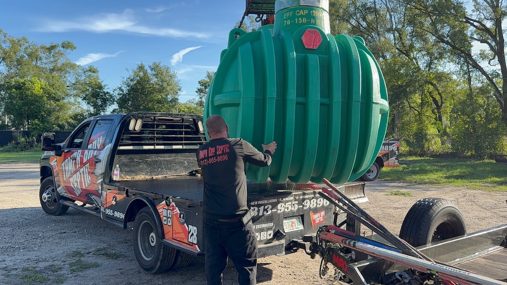 A worker guides a large green plastic septic tank suspended above the back of a flatbed truck.
