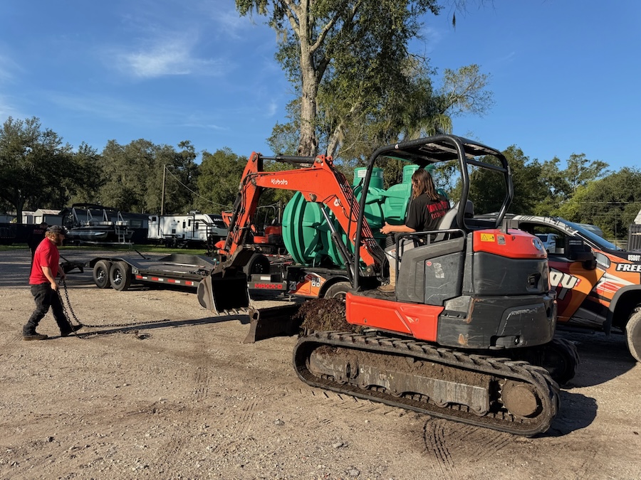 A worker operates an orange mini excavator on a gravel lot near a flatbed trailer loaded with a large green tank while another person handles a tow chain.