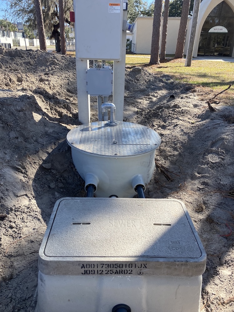 An outdoor sewer infrastructure installation featuring a concrete vault labeled "SEWER," a circular basin with a metal hatch, and an electrical control panel mounted in an excavated dirt area.