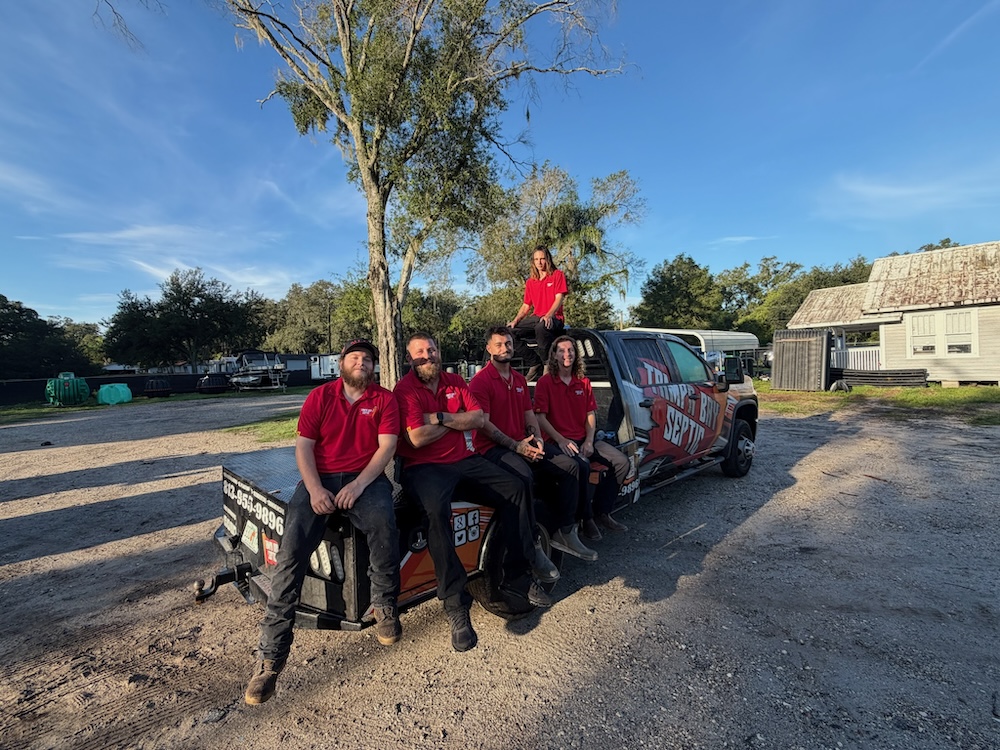A team of six Tampa Bay Septic crew members wearing matching red shirts pose together on and around a branded company truck trailer outdoors.