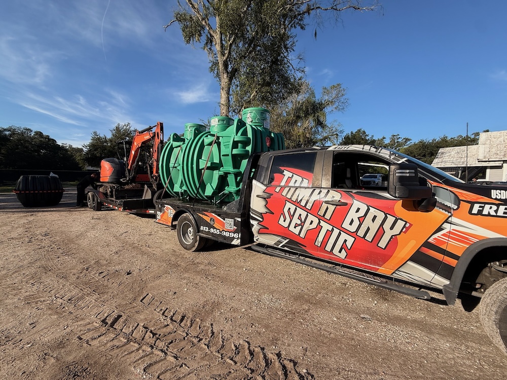 A branded Tampa Bay Septic truck hauls a trailer loaded with large green septic tanks and a mini excavator.