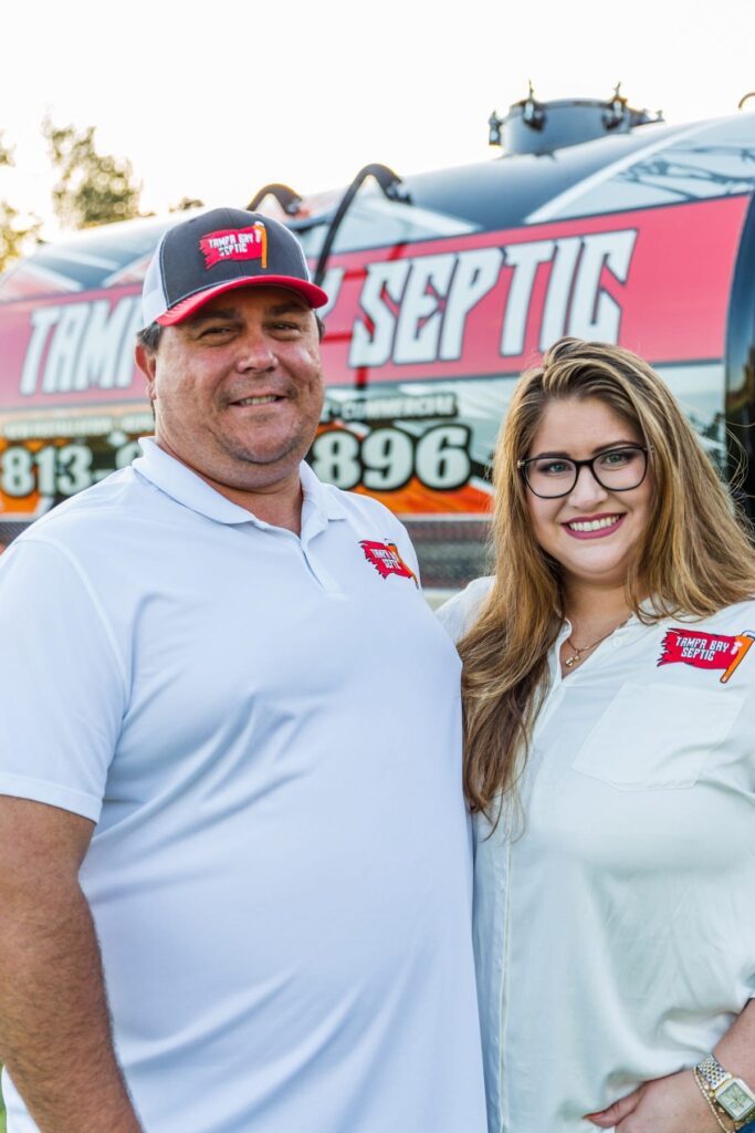 A smiling man and woman in branded white shirts stand together in front of a Tampa Bay Septic service truck.
