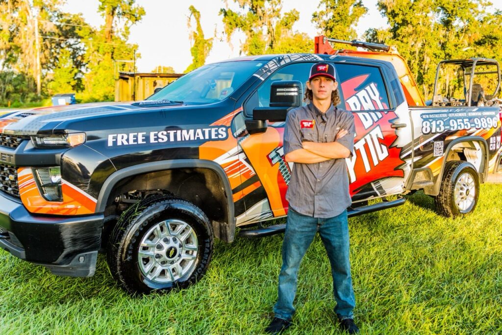 A young man in a grey work shirt stands with arms crossed in front of a custom-wrapped company pickup truck parked on a grassy lawn.
