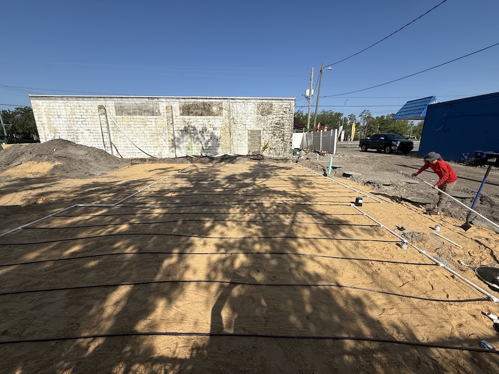 A worker in a red shirt installs a grid of drip irrigation tubing across a cleared sandy lot in front of a weathered masonry building.