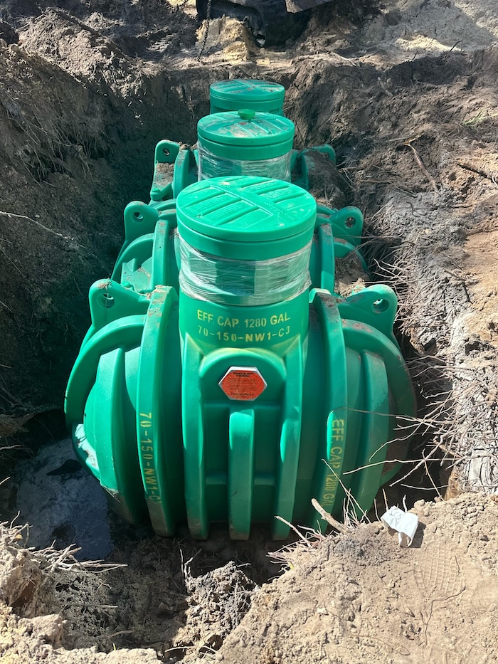 A green polyethylene septic tank with three risers rests inside a newly excavated dirt trench during an installation