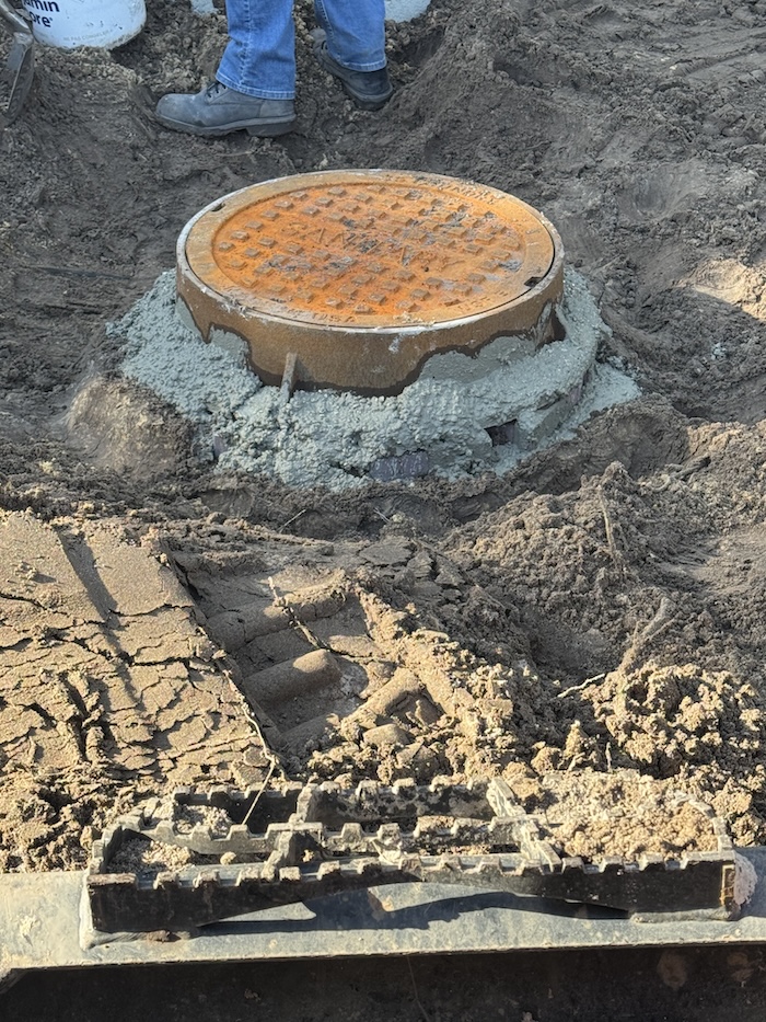 Fresh concrete secures a round cast-iron manhole cover into the ground during a septic tank riser installation.