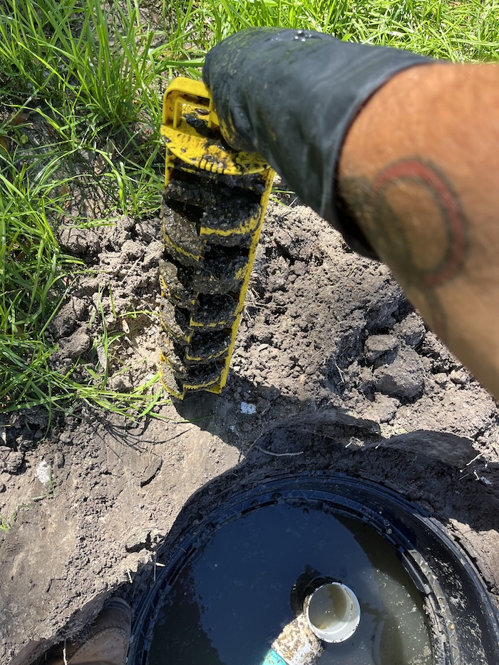 A technician in a black glove pulls a heavily clogged yellow effluent filter out of an open septic tank riser.