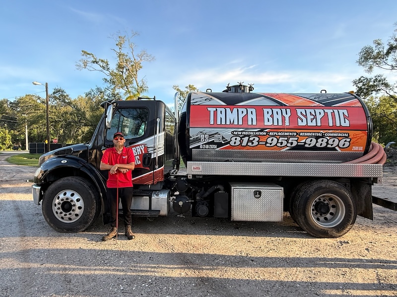 A technician in a red uniform stands beside a branded Tampa Bay Septic vacuum pumper truck on a gravel lot